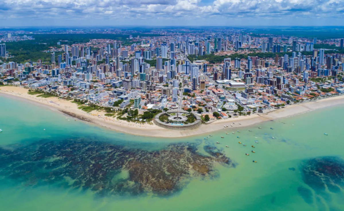 "Vista aérea da orla de João Pessoa, Paraíba, com destaque para os prédios altos, a faixa de areia clara, o mar esverdeado e o letreiro icônico da cidade, representando um destino turístico e urbano em crescimento.